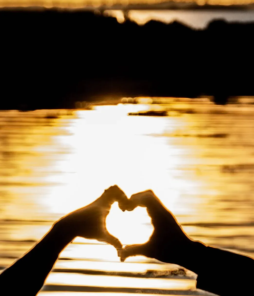 Silhouette de mains formant un cœur devant un coucher de soleil sur l'eau.