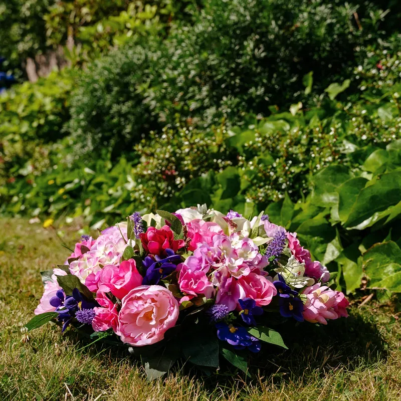 Coussin de fleurs artificielles roses et violettes posé dans l'herbe.